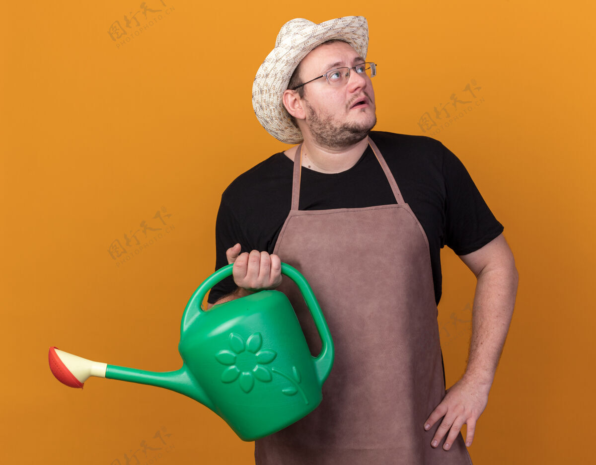 人惊讶地看着身边年轻的男园丁戴着园艺帽拿着浇水罐把手放在臀部隔离在橙色的墙上脸站男