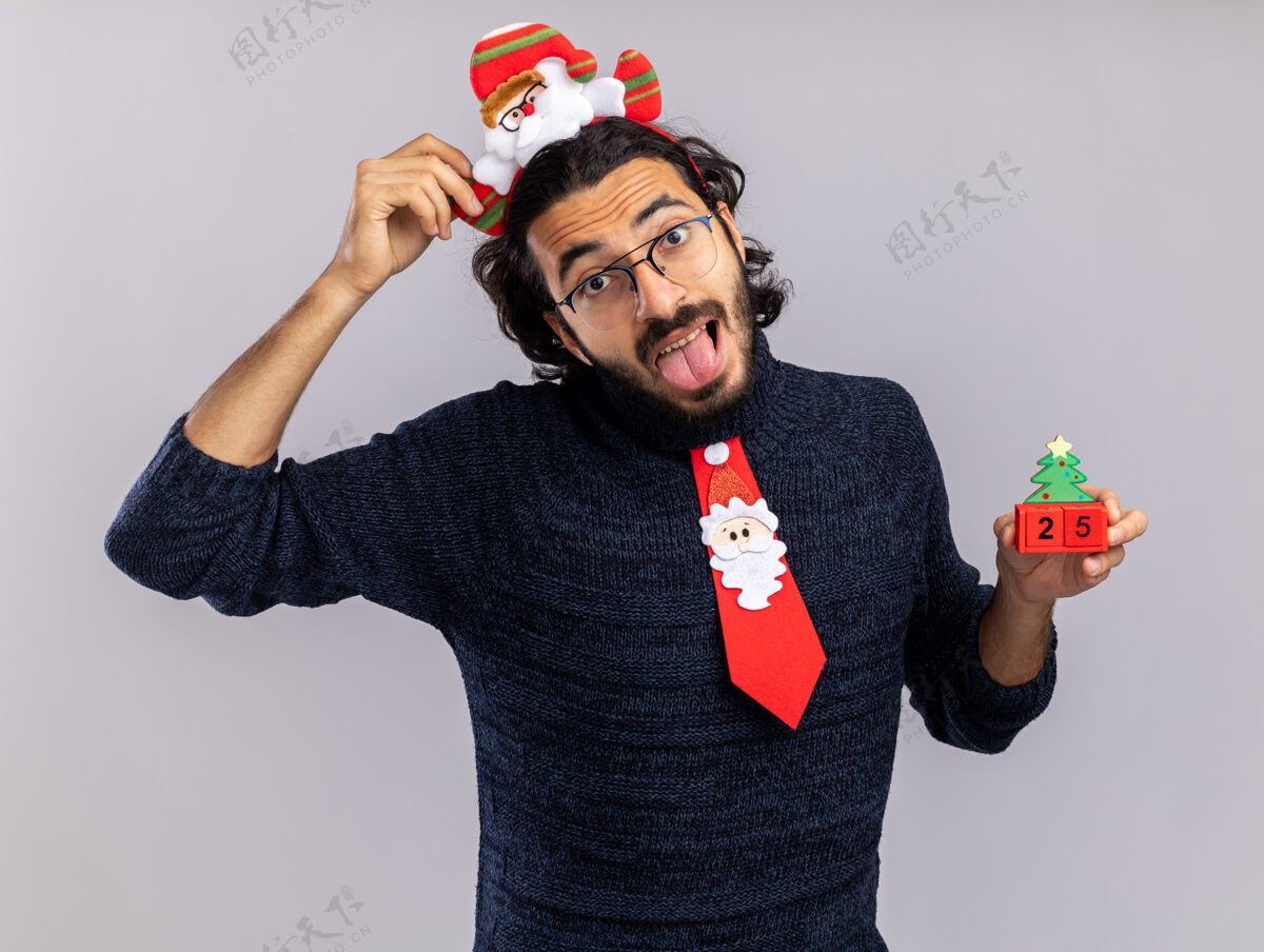 圣诞节快乐的歪头年轻帅哥戴着圣诞领带 戴着发箍 拿着圣诞玩具 在白色背景上露出孤立的舌头玩具倾斜领带