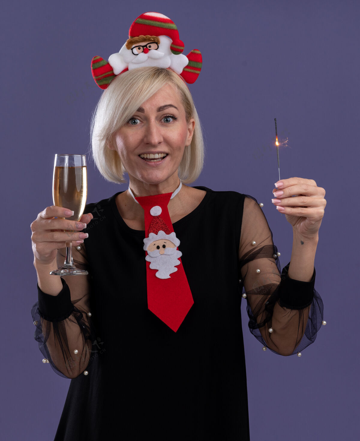 头带快乐的中年金发女人戴着圣诞老人的头带 打着领带 手里拿着节日火花和一杯香槟 看着孤立在紫色背景上的相机领带新年快乐金发