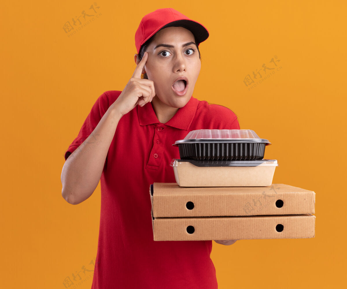 制服令人印象深刻的年轻送货女孩穿着制服 戴着帽子拿着比萨饼盒和食品容器把手指放在隔离在橙色墙上的太阳穴上披萨姿势送货