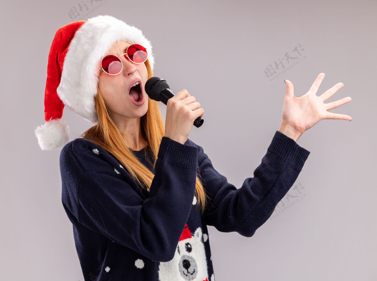 眼镜快乐与闭上眼睛年轻漂亮的女孩戴着圣诞帽和眼镜在麦克风上唱歌传播手隔离在白色背景上麦克风年轻女孩