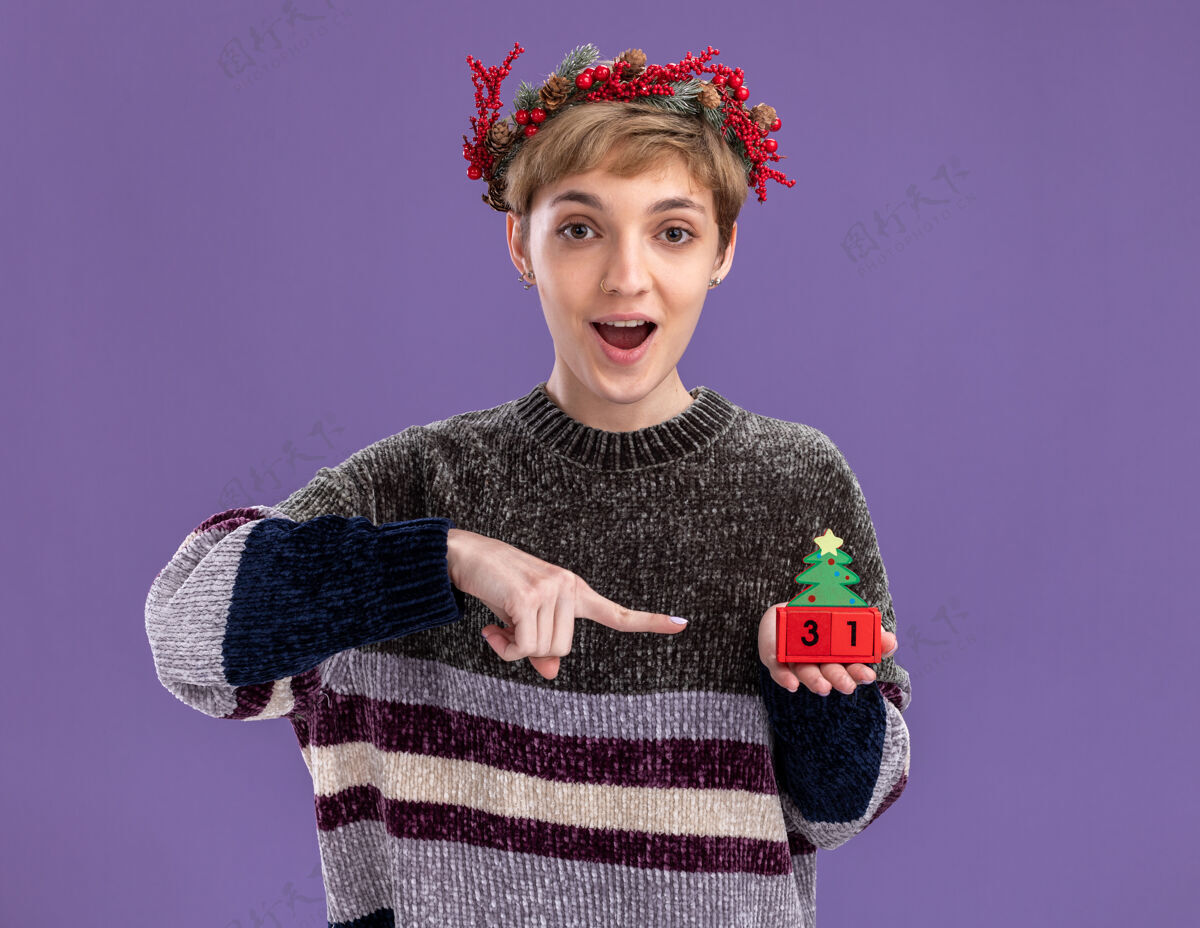 圣诞树给人印象深刻的年轻漂亮女孩戴着圣诞花环拿着圣诞树玩具和日期指着它看着隔离在紫色背景上的相机圣诞快乐年轻紫色