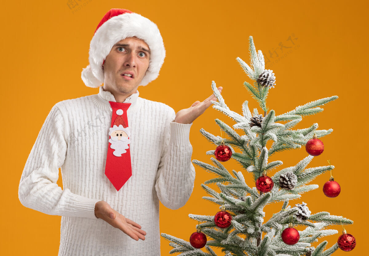 圣诞老人印象深刻的年轻帅哥戴着圣诞帽 打着圣诞老人的领带 站在装饰好的圣诞树旁 用手指着树 看着橙色背景上孤立的摄像头小伙子领带圣诞