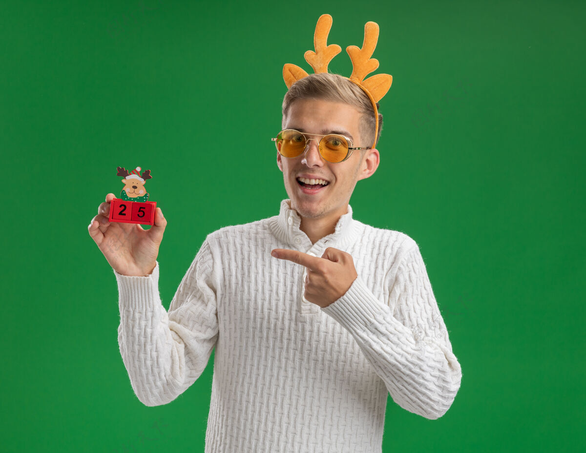 圣诞令人印象深刻的年轻帅哥戴着驯鹿鹿角头带戴着眼镜拿着并指着圣诞树玩具和日期看着隔离在绿色背景上的相机男孩驯鹿圣诞快乐