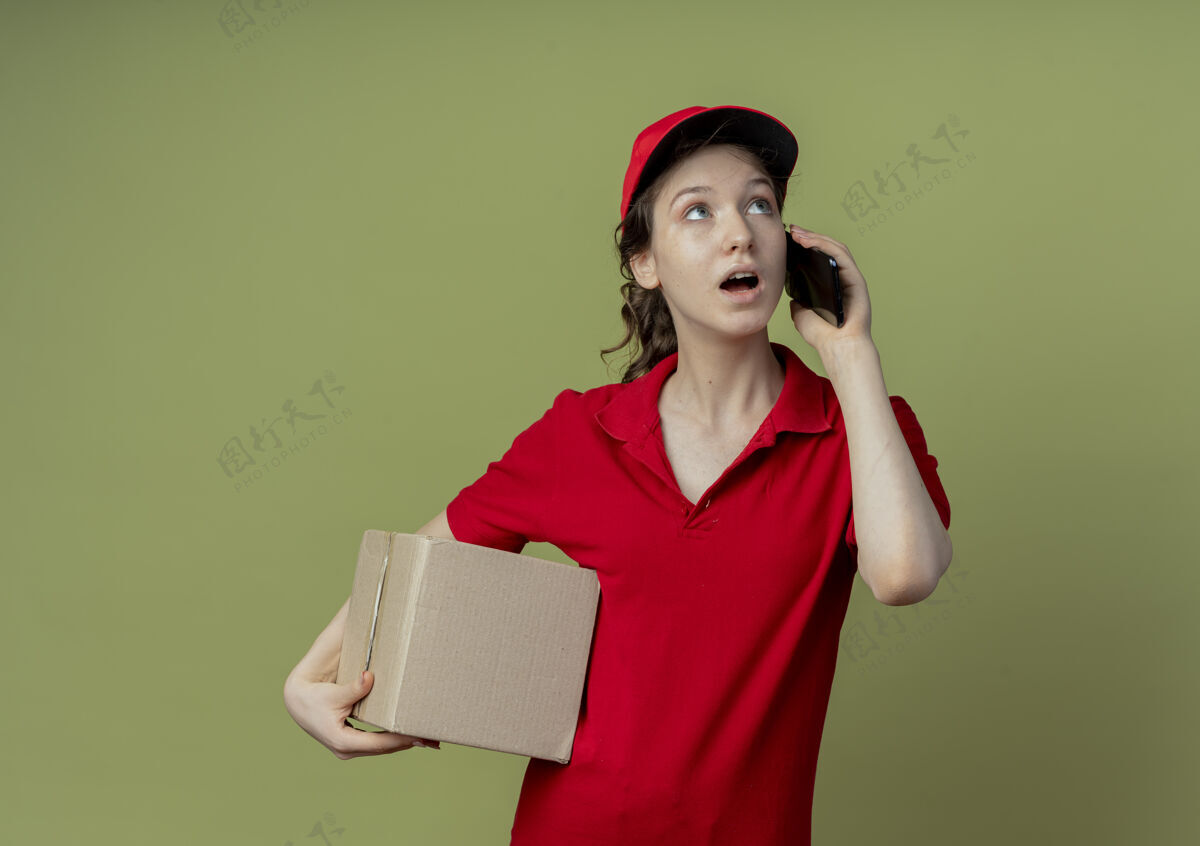 漂亮令人印象深刻的年轻漂亮的送货女孩 穿着红色制服 戴着帽子 抬起头 拿着纸箱 在橄榄绿的背景下 隔着一个复印空间 讲着电话女孩纸箱橄榄色
