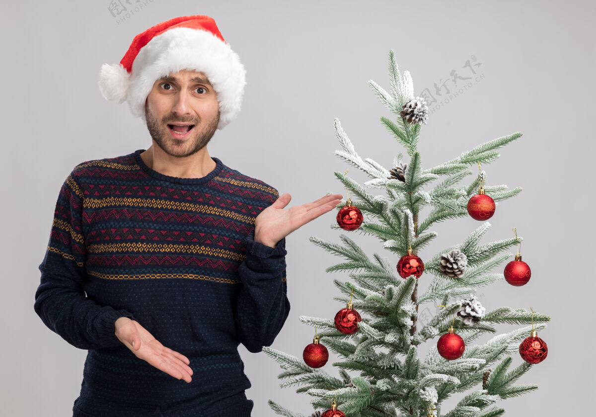 圣诞令人印象深刻的年轻白人男子戴着圣诞帽站在圣诞树旁看着相机用手指着树上孤立的白色背景印象帽子人