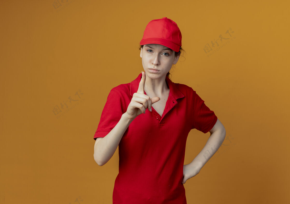 女孩皱着眉头的年轻漂亮的送货女孩 穿着红色制服 戴着帽子 把手放在腰上 举起手指对着隔离在橙色背景上的相机 还有复印空间年轻手帽子