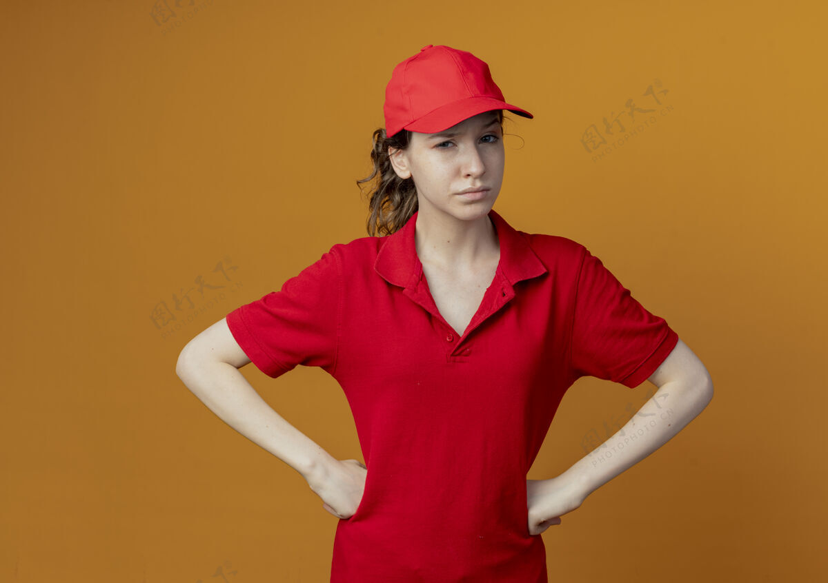 制服可疑的年轻漂亮的送货女孩 穿着红色制服 戴着帽子 手放在腰上 看着隔离在橙色背景上的相机 还有复印空间手年轻怀疑