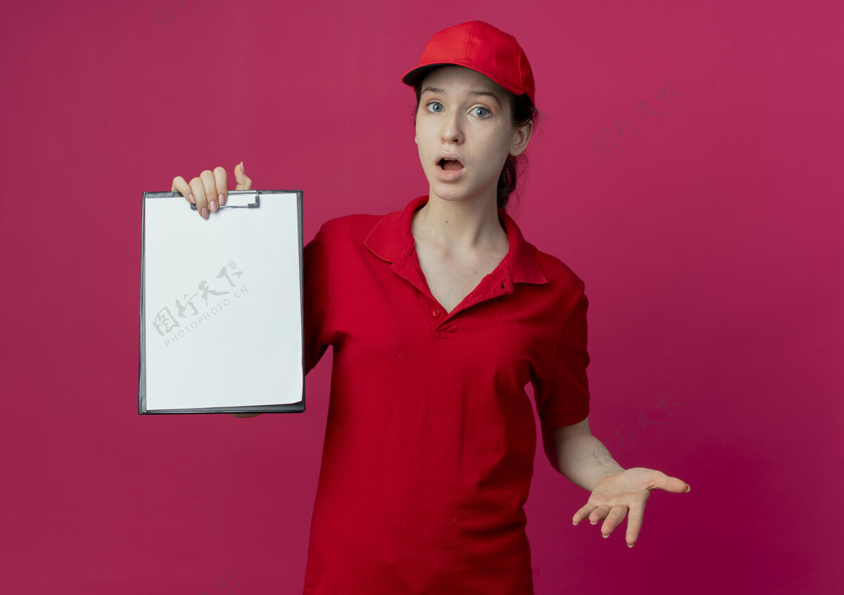 帽子令人印象深刻的年轻漂亮的送货女孩在红色制服和帽子显示剪贴板和空的手孤立在深红色的背景交货显示空