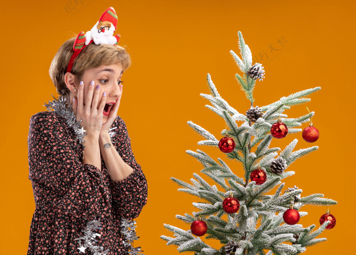 橘子兴奋的年轻漂亮女孩戴着圣诞老人的头带 脖子上戴着金属丝的花环 站在装饰好的圣诞树旁 手放在脸上 在橙色的背景下孤立地往下看手圣诞老人圣诞