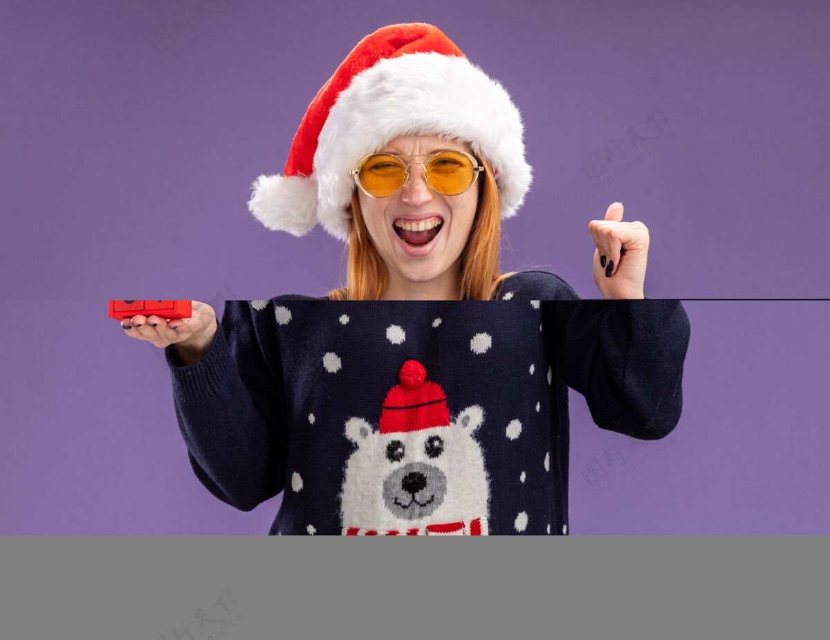 显示兴奋的年轻漂亮女孩穿着圣诞毛衣和帽子戴着眼镜拿着圣诞玩具显示是的手势孤立在紫色背景上眼镜帽子圣诞节