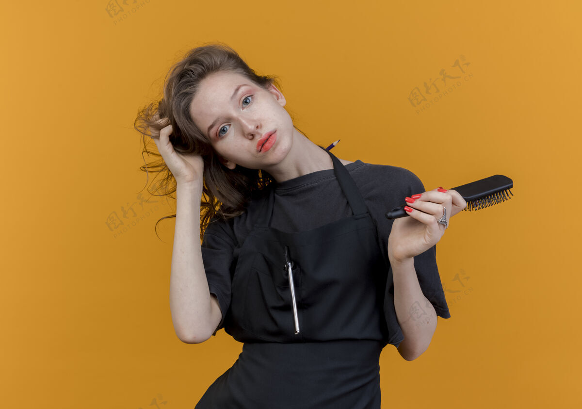 背景年轻的斯拉夫女理发师穿着制服看着相机拿着梳子 抓着头发孤立在橙色背景与复制空间梳子女年轻