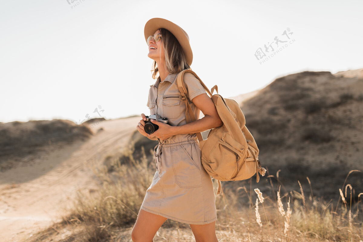 沙漠沙漠中穿着卡其色长裙的迷人时尚年轻女子 在非洲旅行 戴着帽子和背包 用老式相机拍照游客配件背包