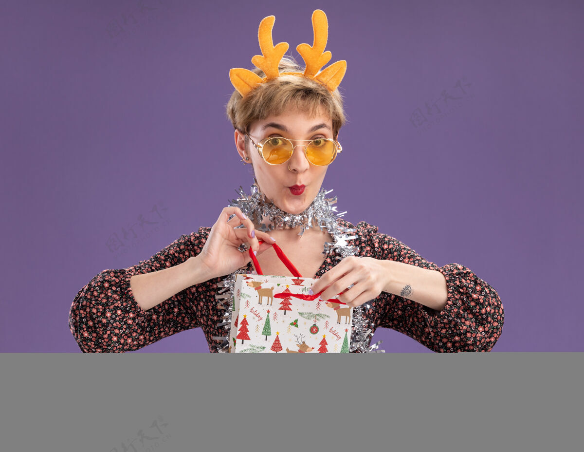 周围好奇的年轻漂亮女孩戴着驯鹿鹿角头带 脖子上戴着金箔花环 戴着眼镜 手里拿着圣诞礼品袋 打开它 看着紫色背景上孤立的相机女孩举行好奇