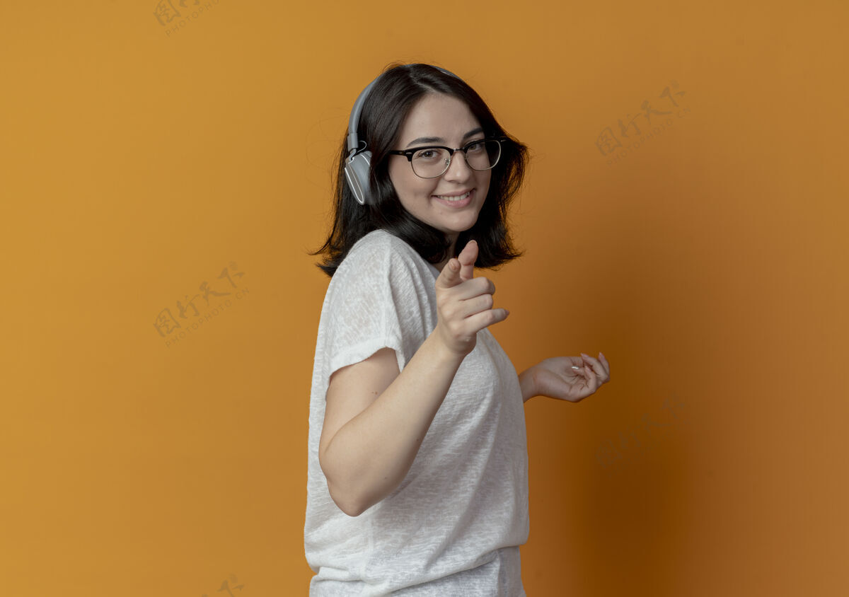 手微笑着的年轻漂亮的白人女孩戴着眼镜和耳机站在侧视图中 手举在空中 指着隔离在橙色背景和复印空间的相机指向橙色个人资料