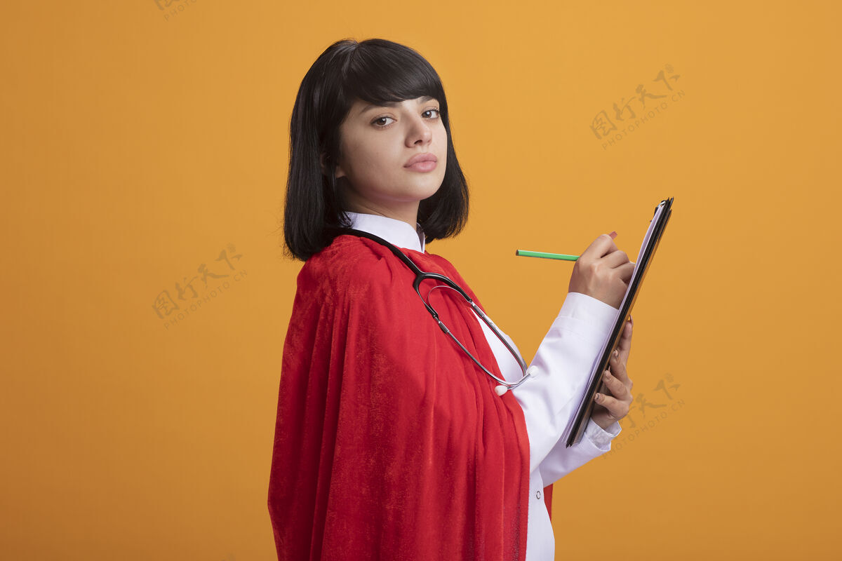 斗篷年轻的超级英雄女孩戴着听诊器 穿着医用长袍和斗篷 在剪贴板上写着什么写作支架年轻人