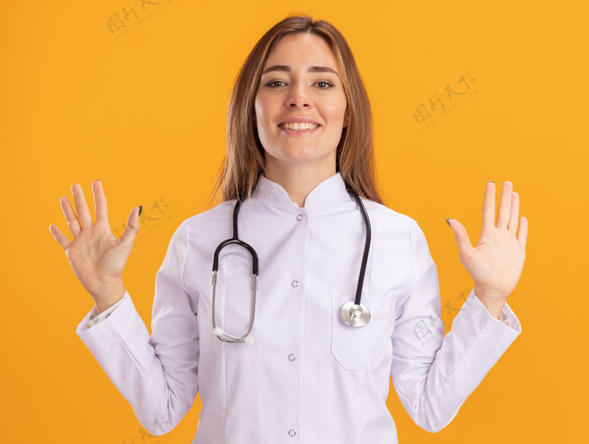 表情面带微笑的年轻女医生穿着医用长袍 听诊器摊开双手隔离在黄色的墙上长袍手人