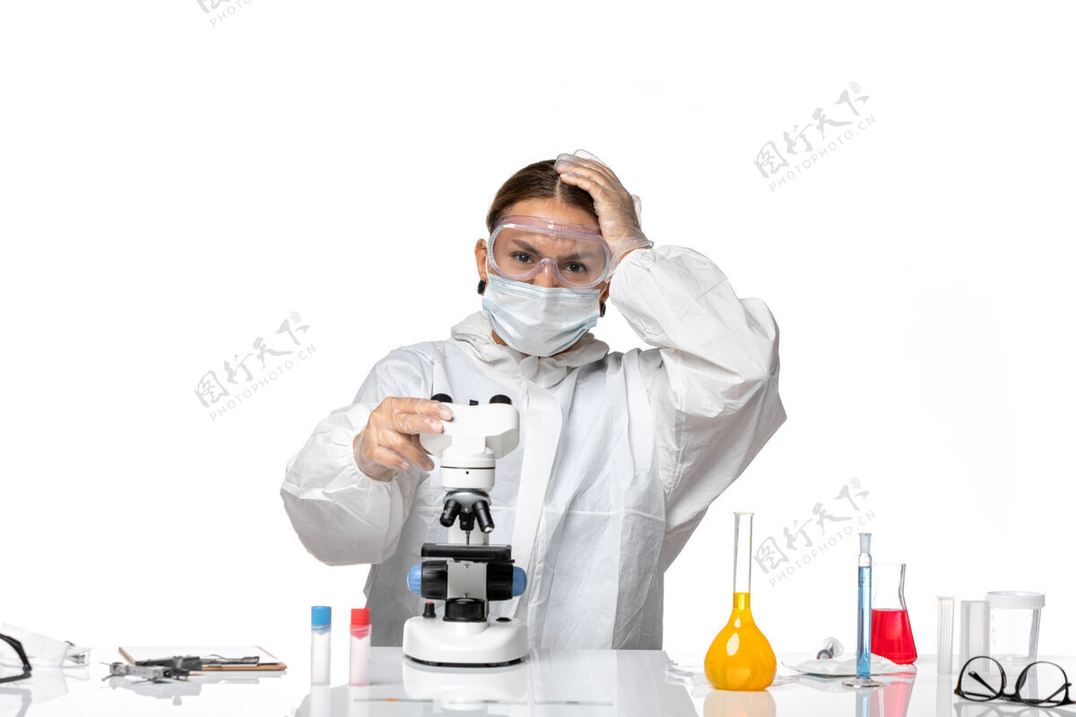 使用前视图女医生穿着特制的西装 戴着口罩 用显微镜观察白色背景病毒冠状病毒大流行实验室外套显微镜口罩