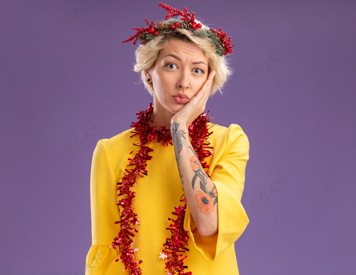 圣诞焦急的金发女郎戴着圣诞花环 脖子上戴着金属丝花环 手放在脸上 看着紫色背景上孤立的摄像机紫色年轻金属丝