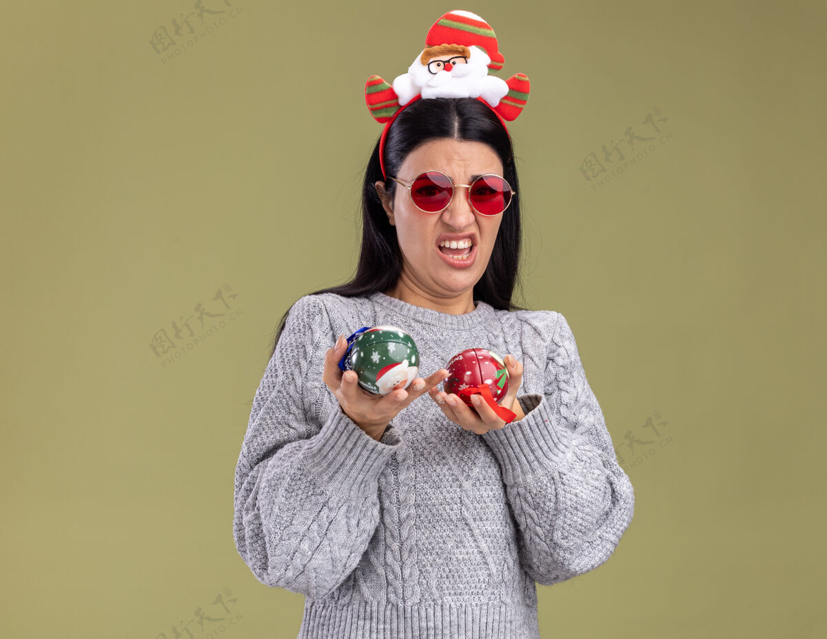 绿色恼怒的年轻白人女孩戴着圣诞老人的头带 戴着眼镜 拿着圣诞饰品 看着橄榄绿背景上的相机举行年轻圣诞