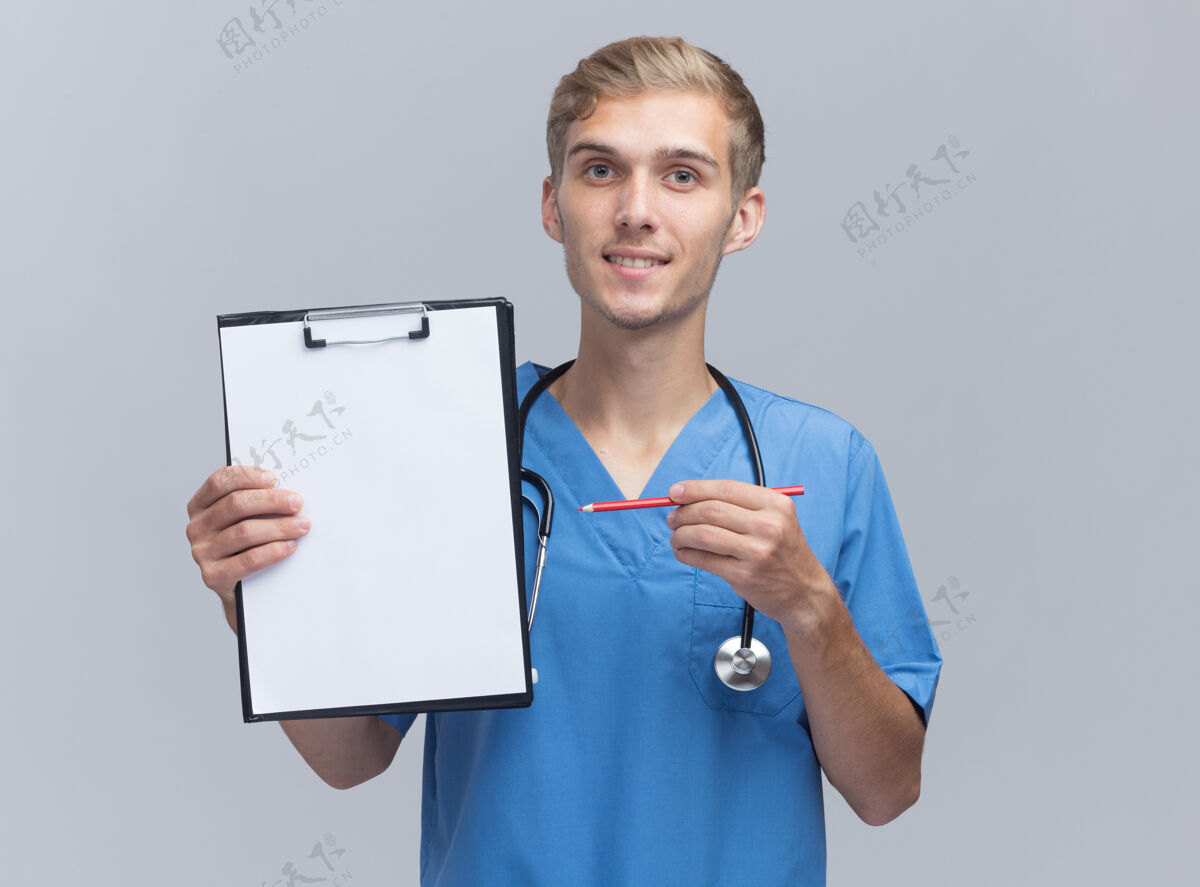 感觉面带微笑的年轻男医生穿着医生制服 手持听诊器 用铅笔指着隔离在白墙上的剪贴板穿着公民点