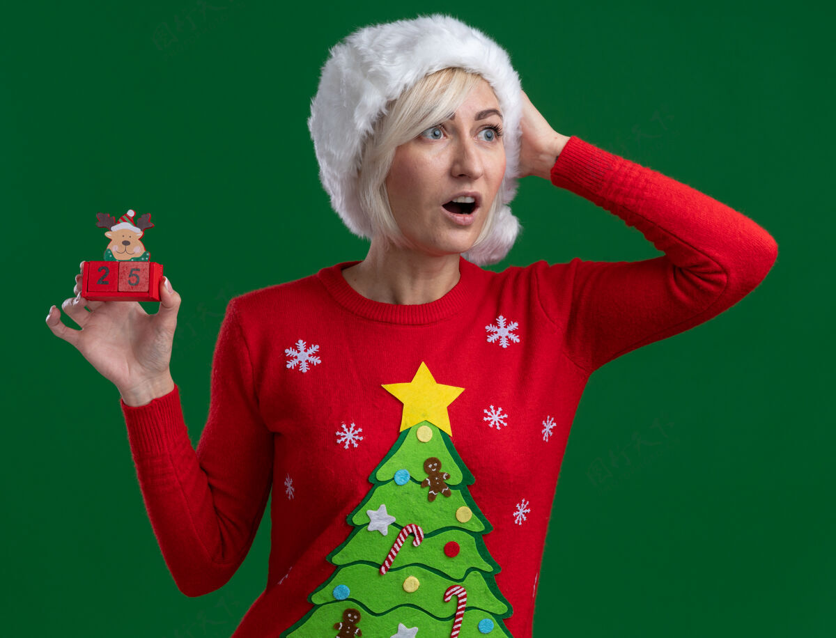 中年令人印象深刻的中年金发女人戴着圣诞帽和毛衣拿着圣诞驯鹿玩具和日期看一边保持手在头上绿色背景隔离举行印象驯鹿