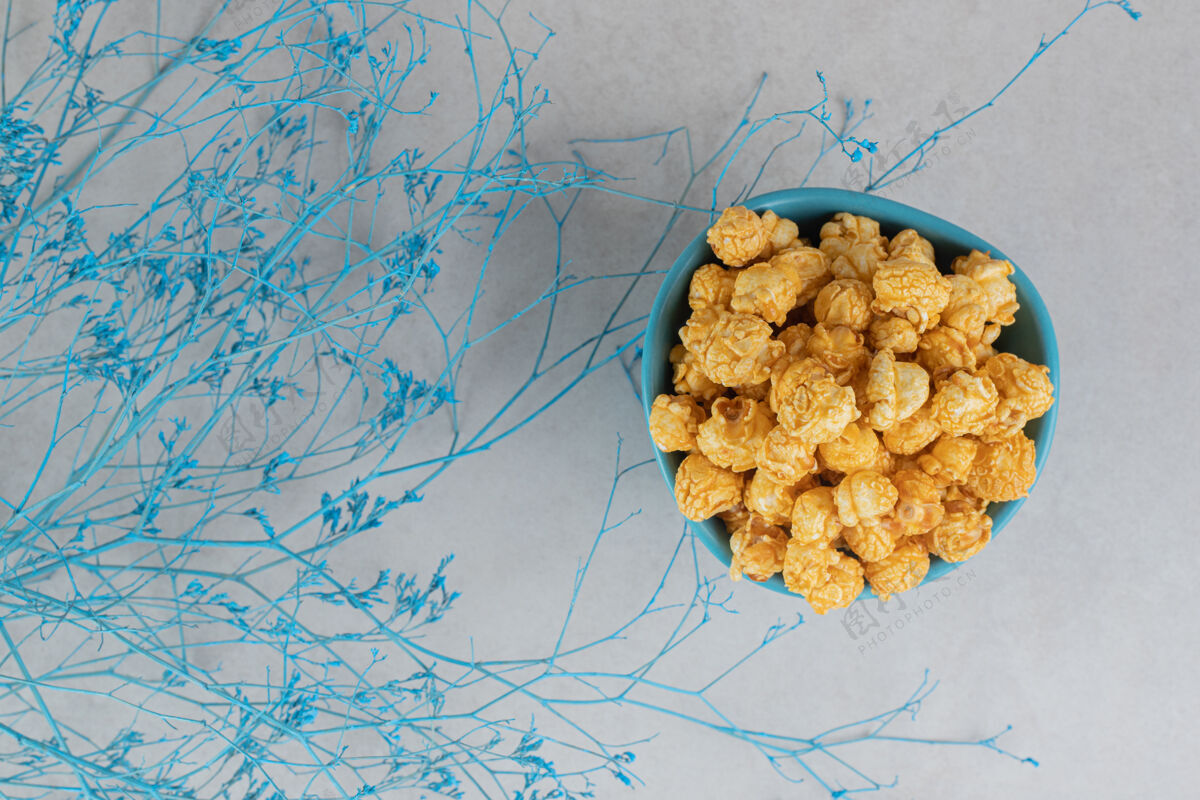 焦糖一碗涂着焦糖的爆米花 放在大理石桌上 蓝色的树枝环绕着它树枝爆米花零食