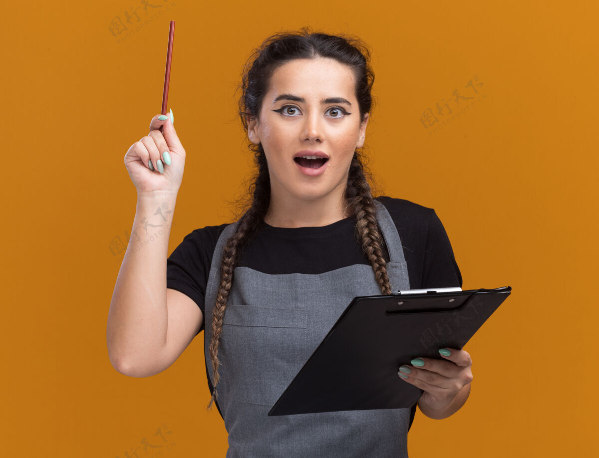制服印象深刻的年轻女理发师在制服举行剪贴板和提高铅笔隔离在橙色的墙上女性拿着脸