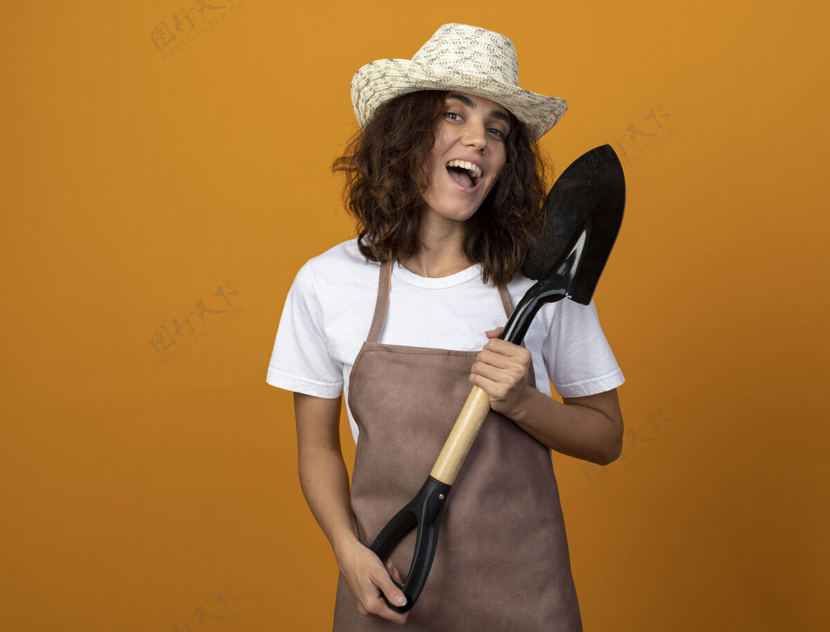 女人快乐的年轻女园丁穿着制服 戴着园艺帽 拿着铲子黑桃穿制服