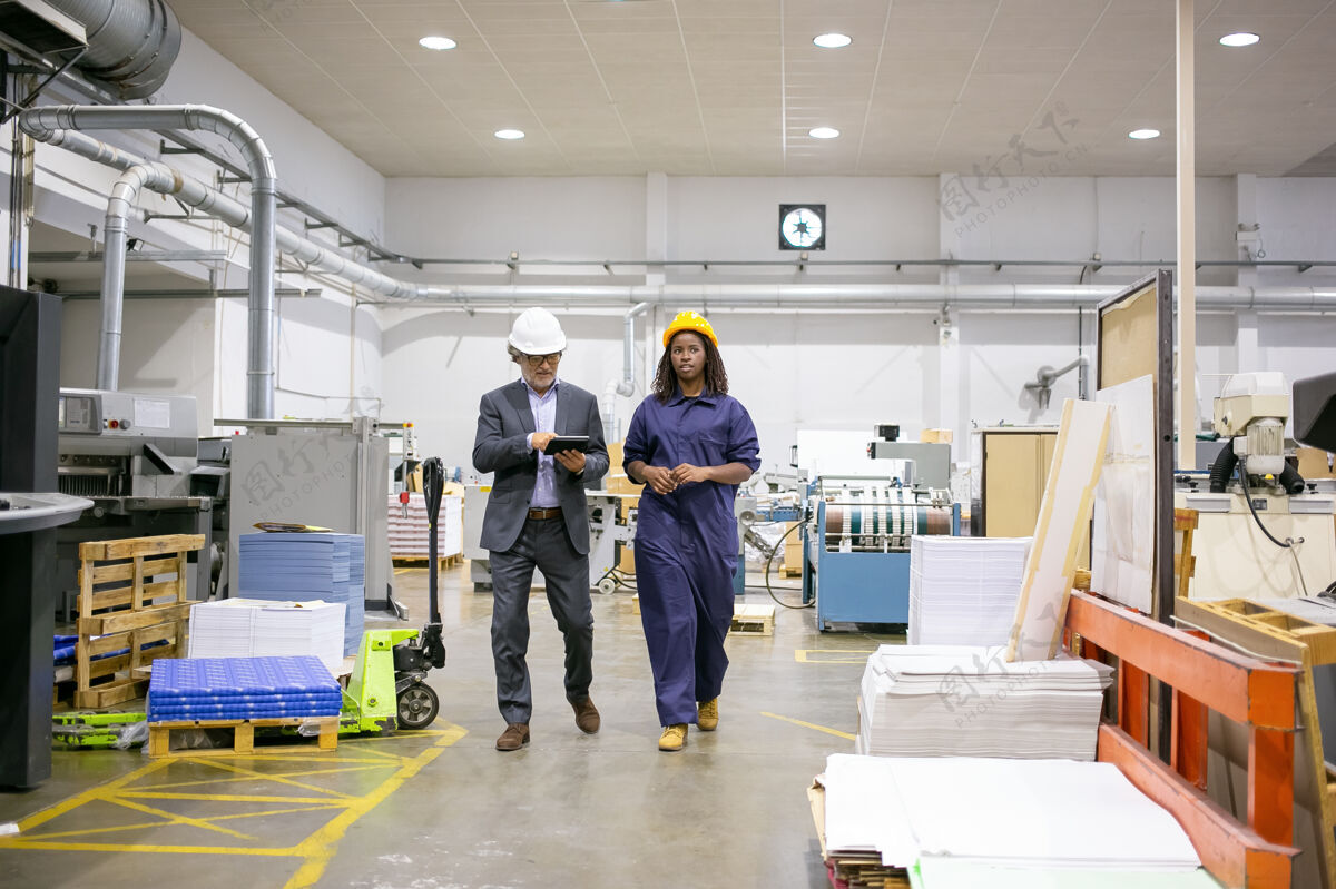 工作严肃的男检查员和戴着安全帽的女工厂员工走在车间地板上说话 男人在用平板电脑男性安全工程