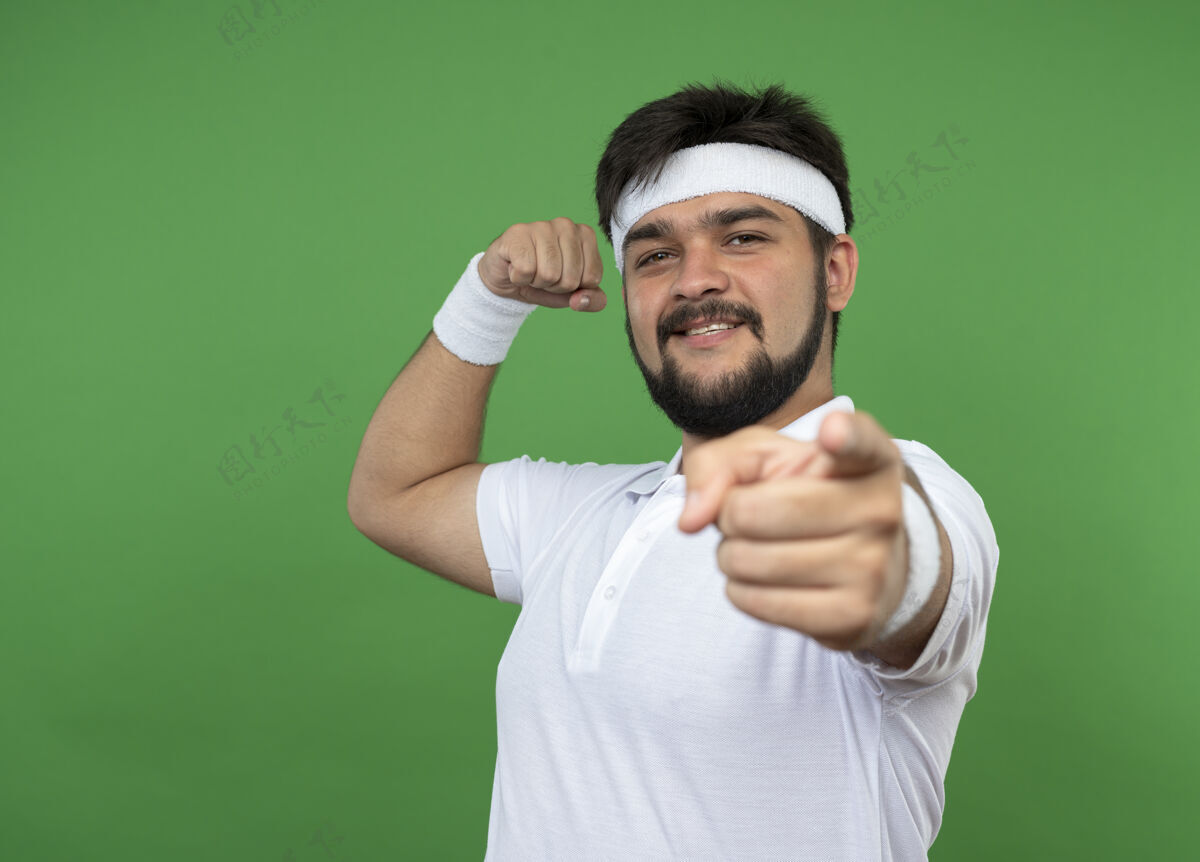 强壮微笑的年轻人戴着头带和腕带显示出强烈的运动姿态点隔离在绿色墙壁与复制空间手势头带秀