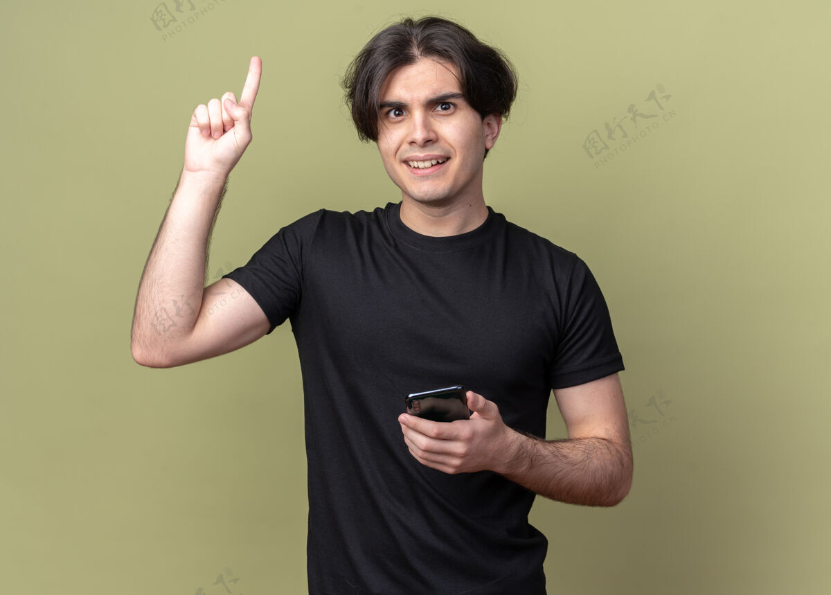 点印象深刻的年轻帅哥穿着黑色t恤手持电话点在橄榄绿的墙上孤立起来穿着持有英俊