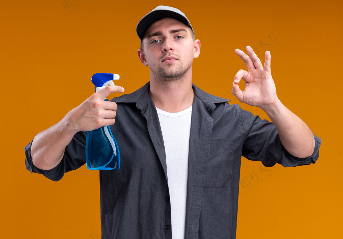 好自信的年轻帅气的清洁工 穿着t恤 戴着帽子 拿着喷雾瓶 在橙色的墙上表现出良好的姿态男人清洁帽子