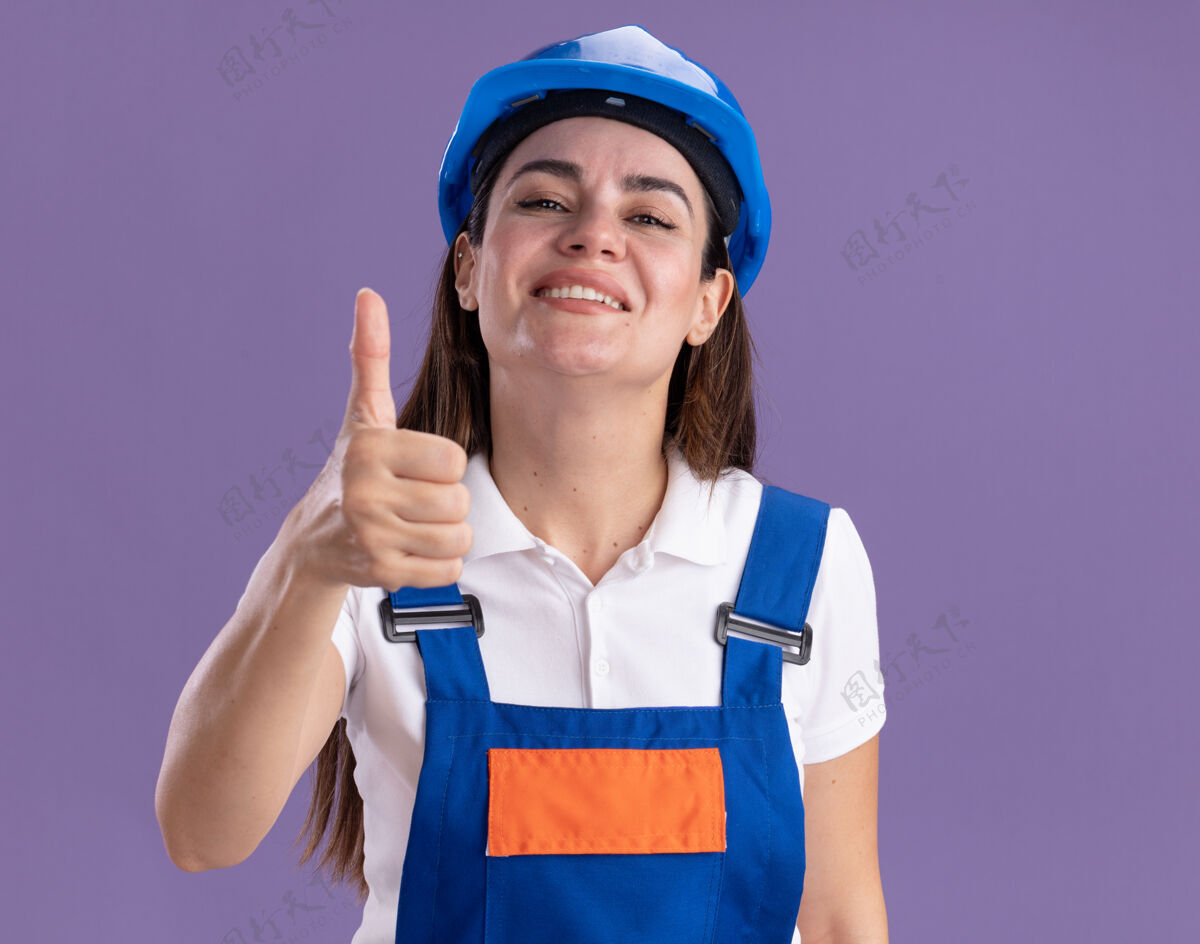 年轻身着制服的年轻建筑工人微笑着 在紫色的墙上孤立地竖起大拇指表演女人微笑
