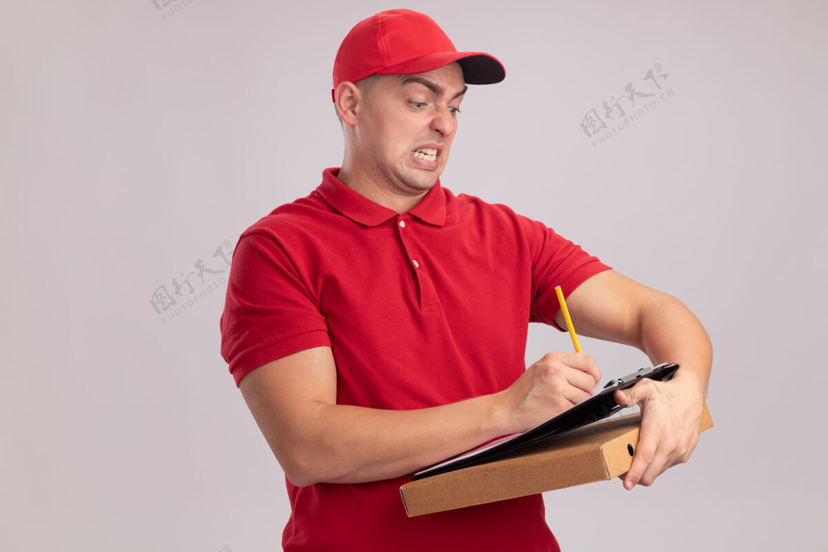 帽子愤怒的年轻送货员穿着制服 戴着帽子 拿着比萨饼盒 在白墙上的剪贴板上写着什么男人什么的戴着