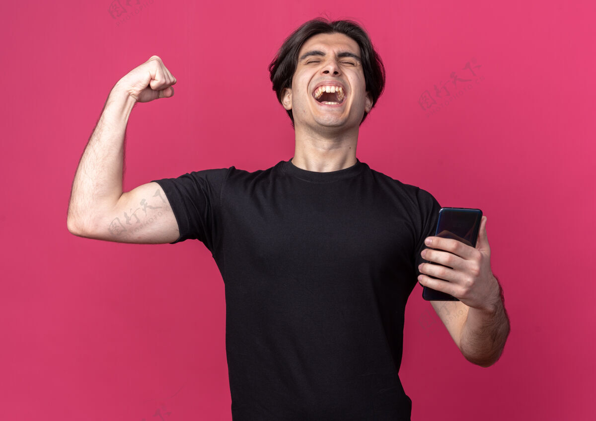 年轻兴奋的闭上眼睛年轻帅哥穿着黑色t恤手持手机表示是的手势孤立在粉红色的墙上兴奋电话举行