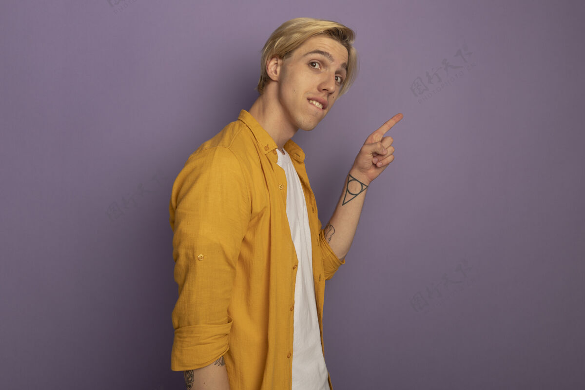 年轻高兴的年轻金发男子穿着黄色t恤点在一边孤立的紫色与复制空间金发男请穿