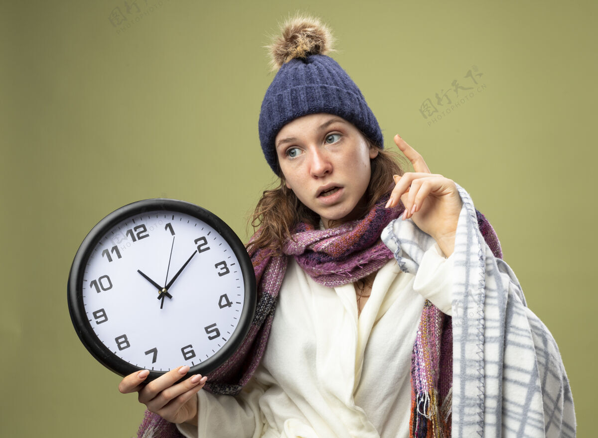 时钟印象深刻的年轻生病的女孩一边看一边穿着白色长袍 戴着冬天的帽子 拿着围巾的挂钟包着格子呢的点在橄榄绿上孤立起来格子举行穿