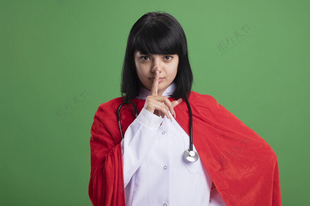 听诊器严格的年轻超级英雄女孩戴着听诊器与医疗袍和斗篷显示沉默的姿态隔离在绿色手势年轻长袍