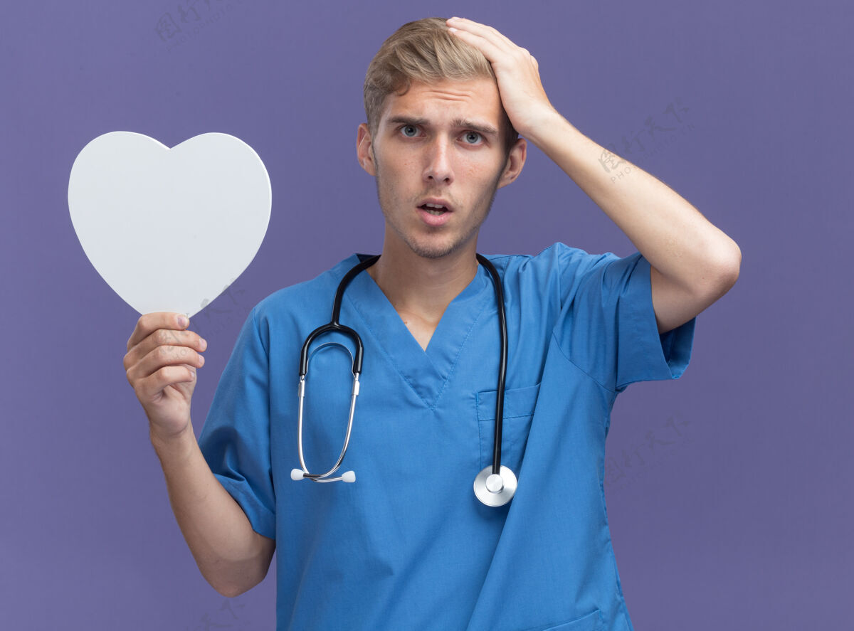 年轻遗憾的是 年轻的男医生穿着医生制服 手持听诊器 手持心形盒子 手放在隔离在蓝色墙上的头上制服男性头