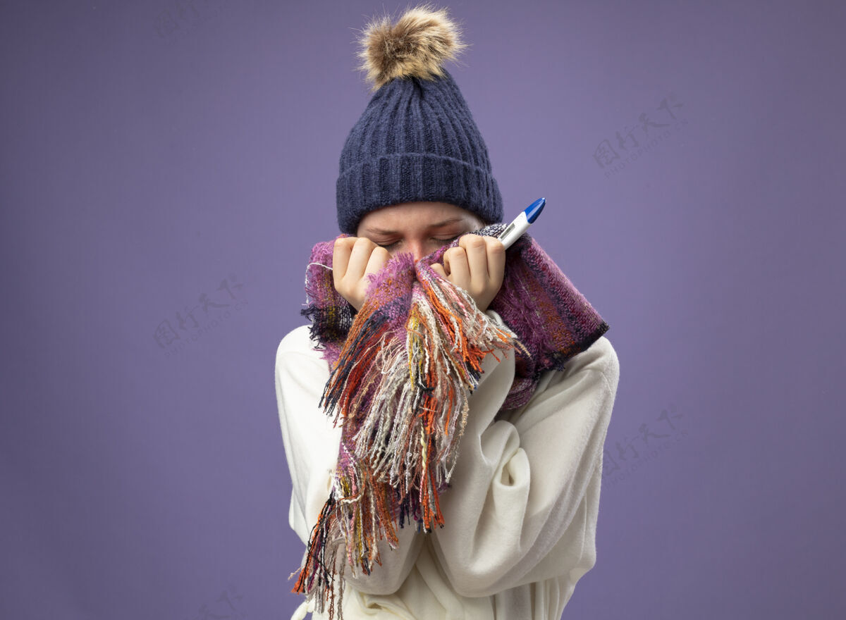 眼睛年轻的病女孩闭着眼睛 穿着白色长袍 戴着冬天的帽子 戴着围巾 拿着温度计 脸上戴着紫色的围巾体温计帽子年轻