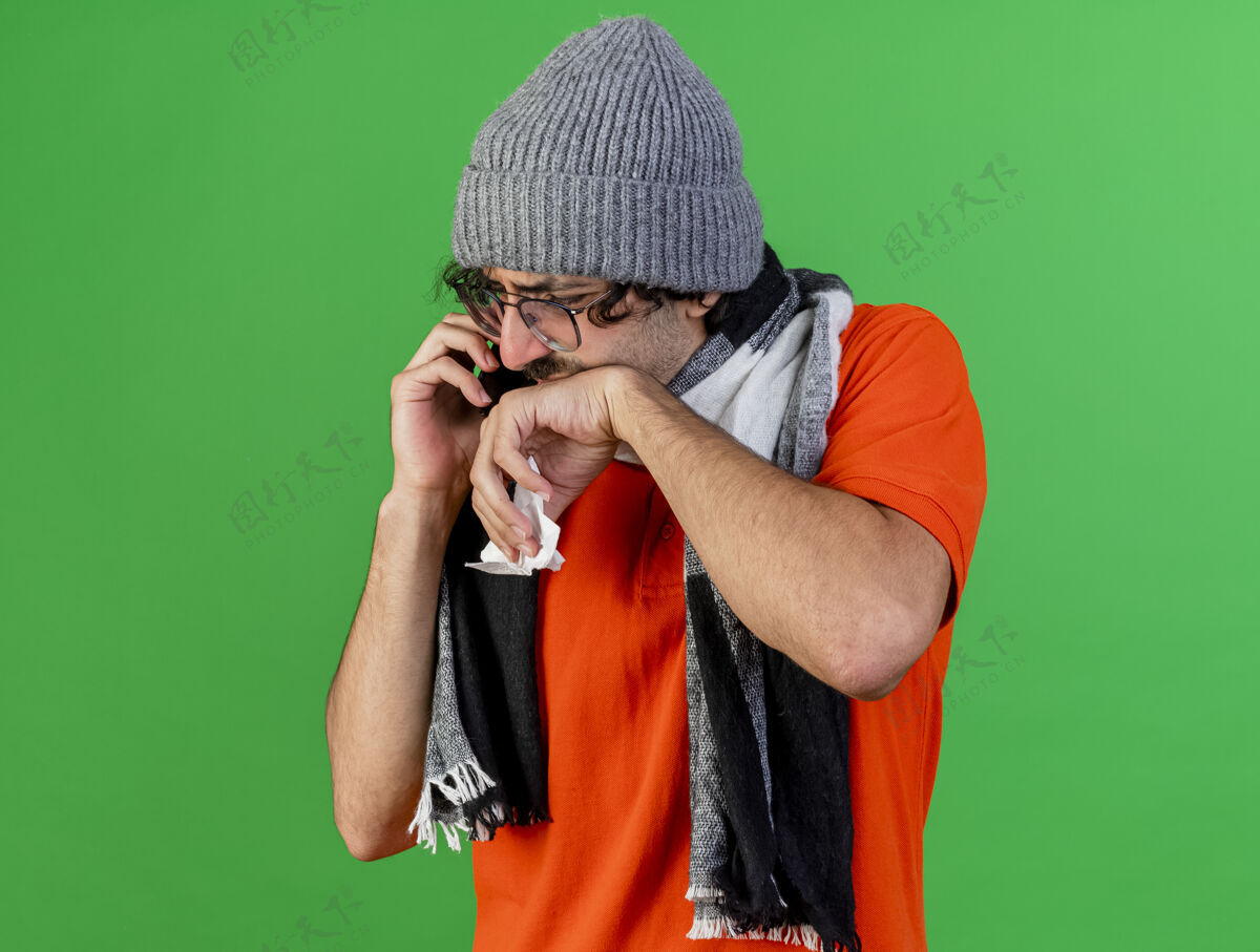 人戴着眼镜 戴着冬天的帽子 戴着围巾 拿着餐巾 手放在嘴上 孤独地看着绿色墙壁 留着复印空间的疼痛的年轻人冬天疾病人