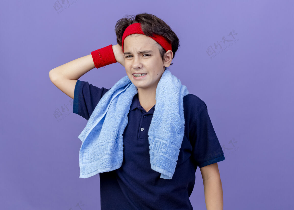 站疼痛的年轻英俊的运动男孩戴着头带和护腕 脖子上戴着牙套和毛巾 隔离在紫色的墙上市民腕带戴