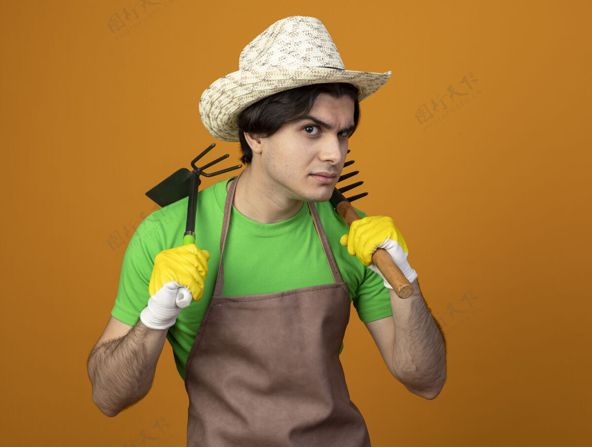 磨损可疑的年轻男园丁穿着制服戴着园艺帽戴着手套拿着锄头耙肩上的耙子隔离在橘子上橙色可疑锄头