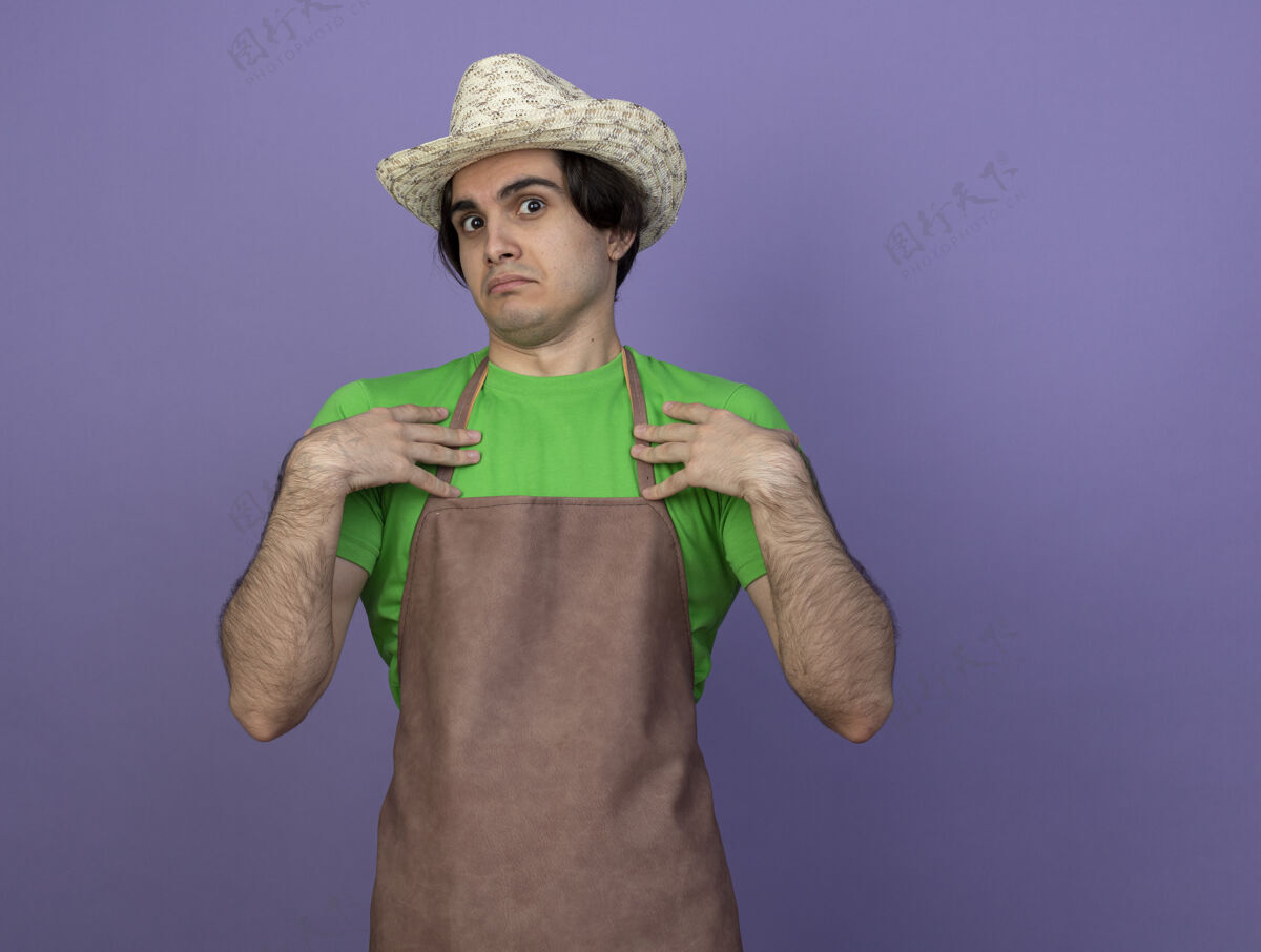 紫色困惑的年轻男园丁穿着制服戴着园艺帽把手放在自己身上困惑园艺帽子