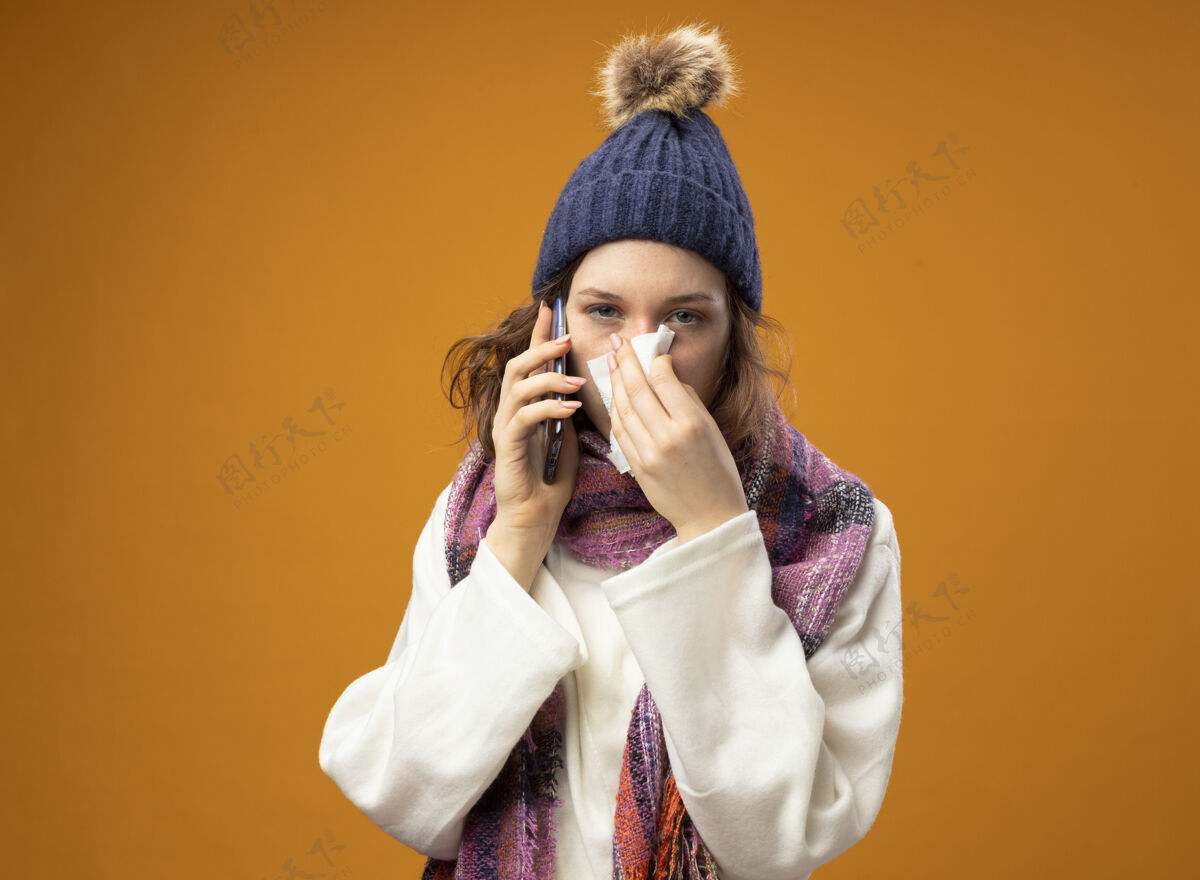围巾年轻的生病的女孩穿着白色长袍 戴着带围巾的冬帽 直直地向前看 一边讲电话一边用橘黄色隔离的餐巾擦鼻子帽子穿餐巾