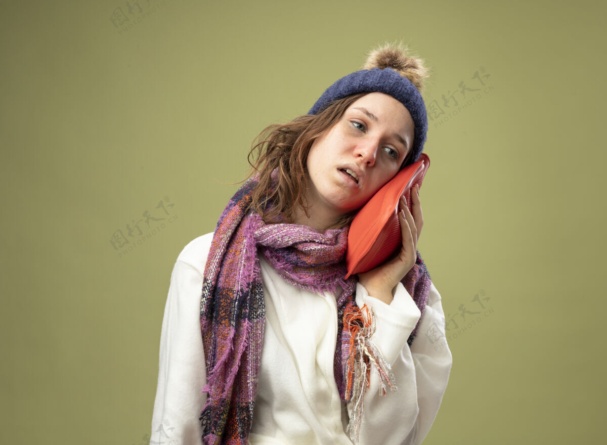 脸颊虚弱的生病的年轻女孩穿着白色长袍 戴着冬天的帽子 戴着围巾 把热水袋放在脸颊上 隔离在橄榄绿上水虚弱绿