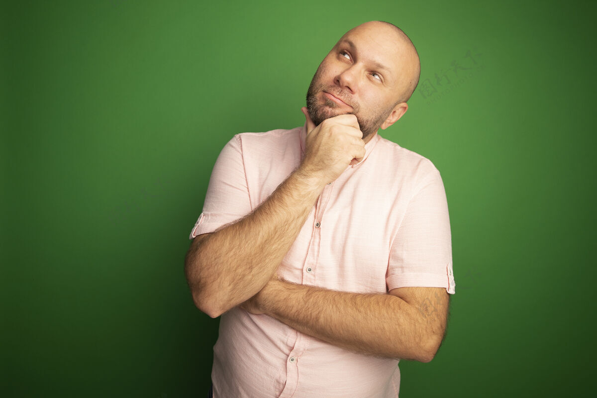中年印象深刻的边看边中年秃头男子穿着粉红色的t恤抓住下巴隔离在绿色的墙上秃头抓住磨损
