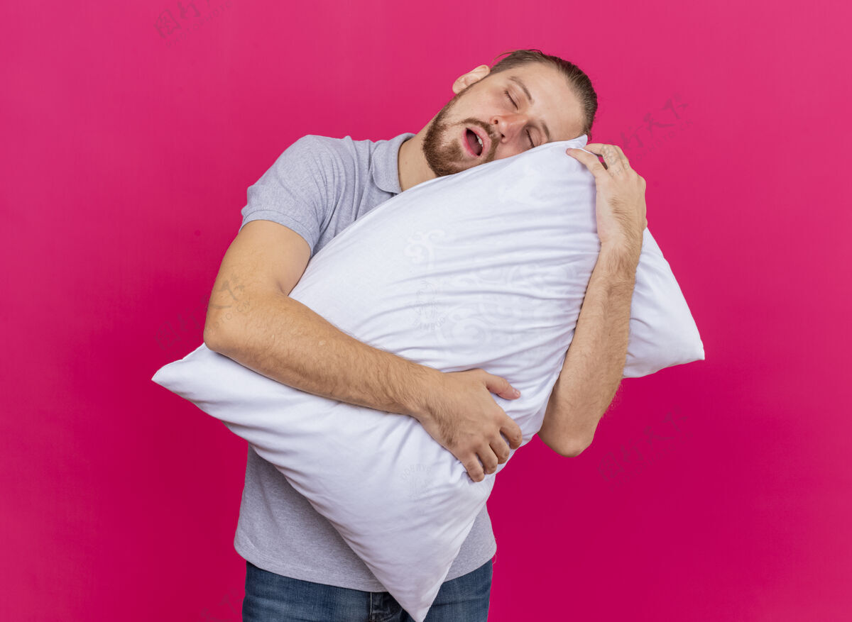 站立疲惫的年轻英俊的斯拉夫病夫抱着枕头把头枕在上面 闭着眼睛孤立在粉红色的墙上衣服眼睛疲倦
