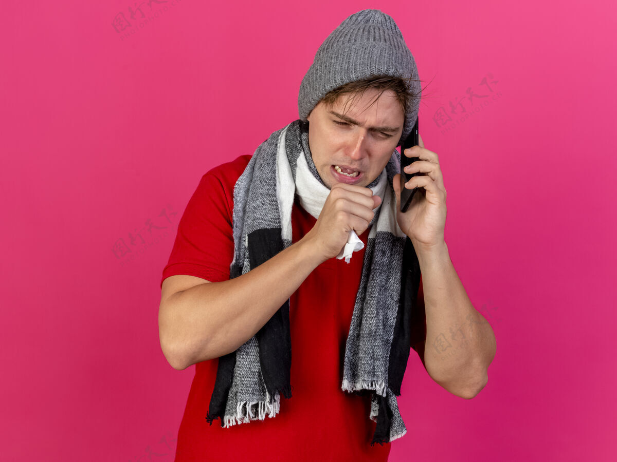 人愁眉不展的年轻英俊的金发病男子戴着冬天的帽子和围巾讲电话拿着餐巾纸把手放在嘴边咳嗽看着隔离在粉红色墙上的复制空间围巾餐巾帅哥